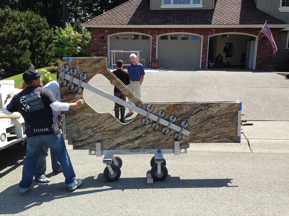 Two workers move a large plywood ramp on a wheeled cart in a driveway in front of a house.