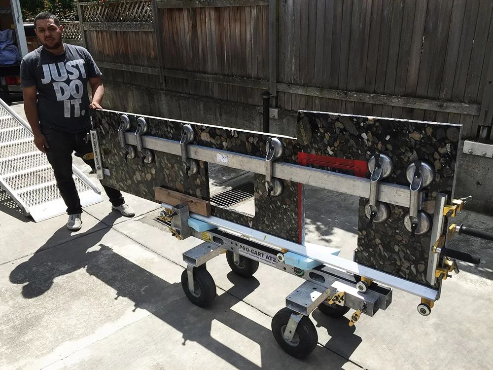 Man standing beside a long metal cart with four wheels in a driveway, near a wooden fence.