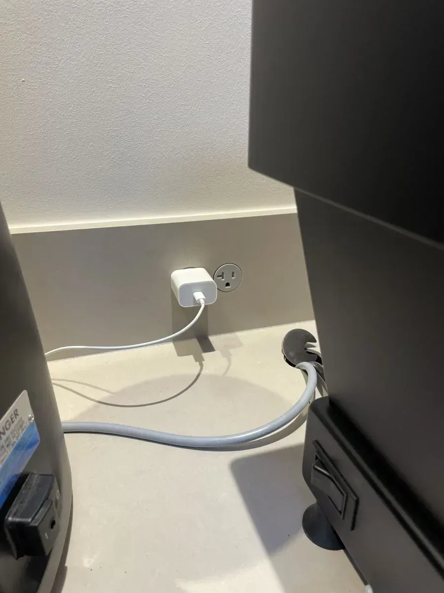 White power adapter plugged into an outlet, with a gray cable across a floor beside black furniture.