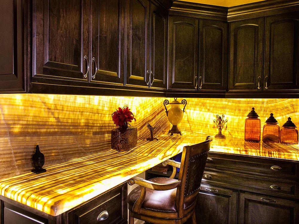 Warmly lit dark wood kitchen corner with glowing amber backsplash, countertop decor, and a chair at the desk.