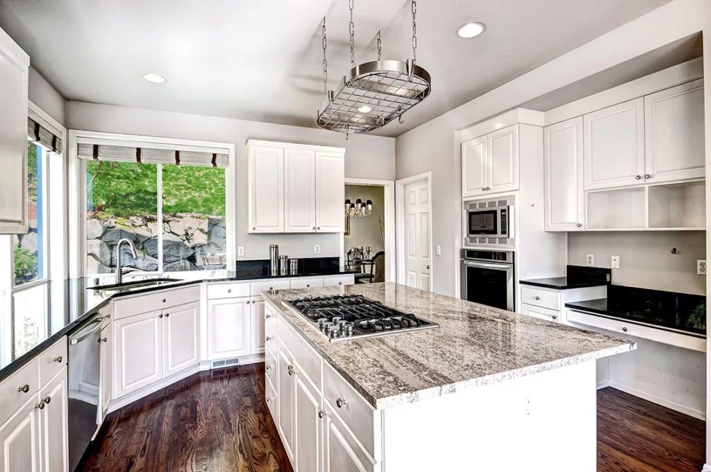 Bright white kitchen with granite island, stainless appliances, and large window overlooking greenery