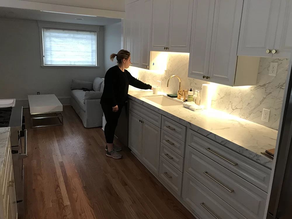 Person standing at a lit kitchen counter in a bright open-plan room with wood floors.