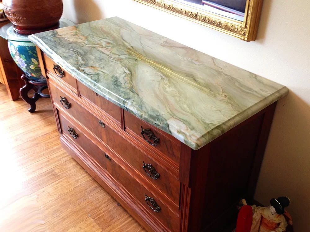 Wooden dresser with a green marble top in a sunlit room, beside a decorative vase and framed wall art.
