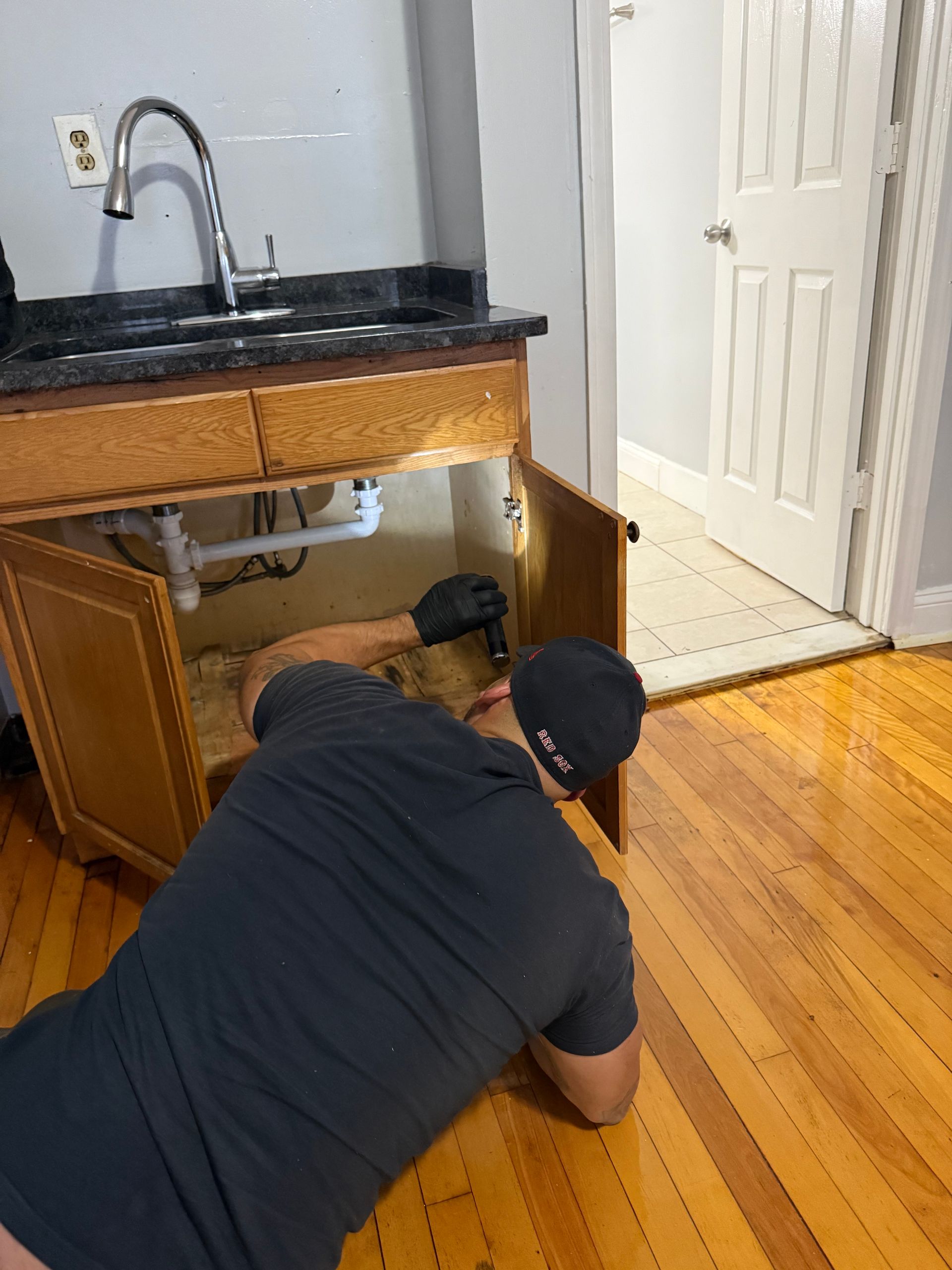 Person in black shirt, cap, gloves working on plumbing under a kitchen sink.