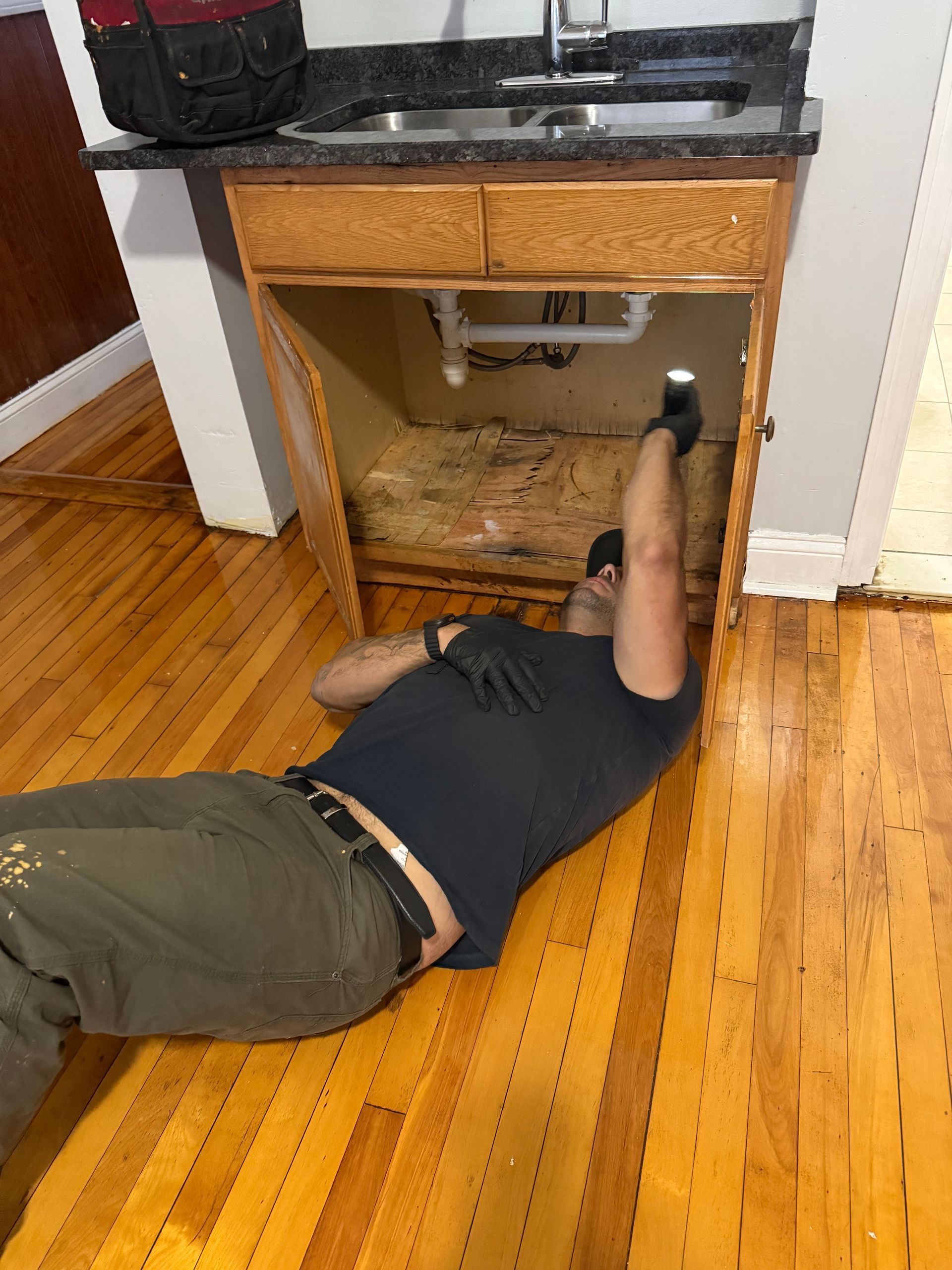 Man working under a kitchen sink. He’s wearing a dark shirt and pants. The cabinet is open. Hardwood floor.