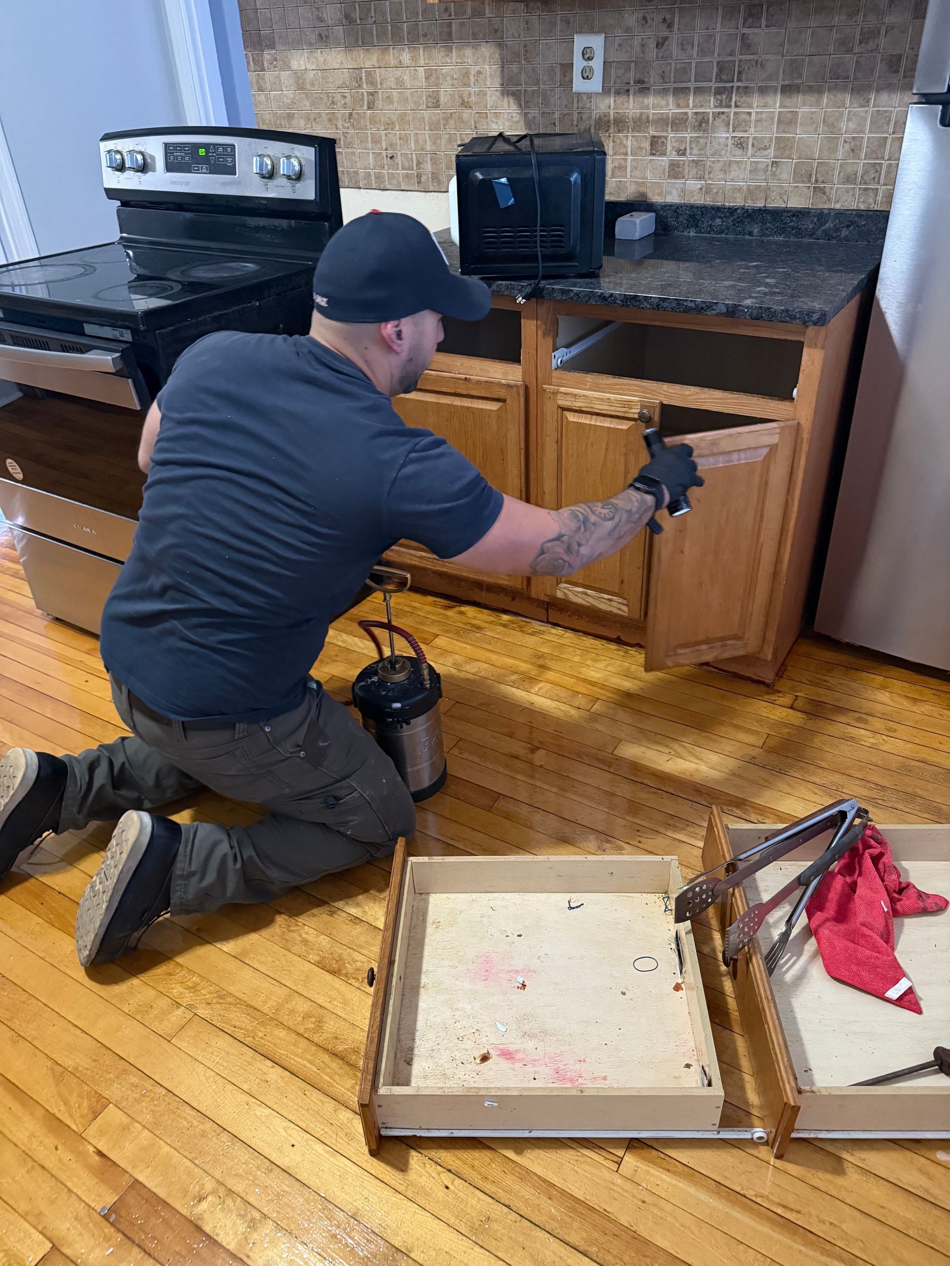 Person kneeling, spraying insecticide inside kitchen cabinet. Stove and drawers visible on hardwood floor.
