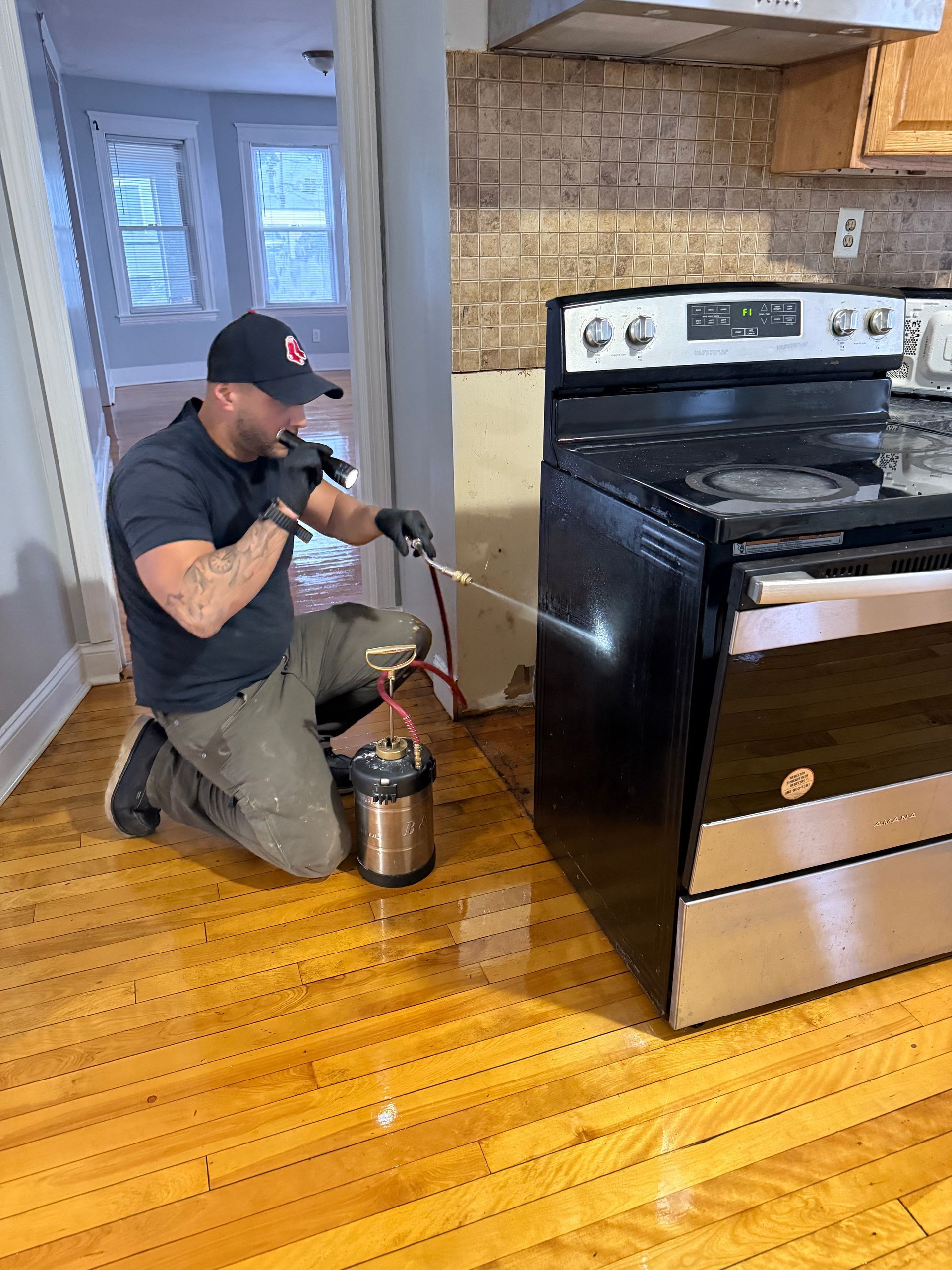 Person in gloves spraying insecticide near a black oven in a kitchen with wood flooring.