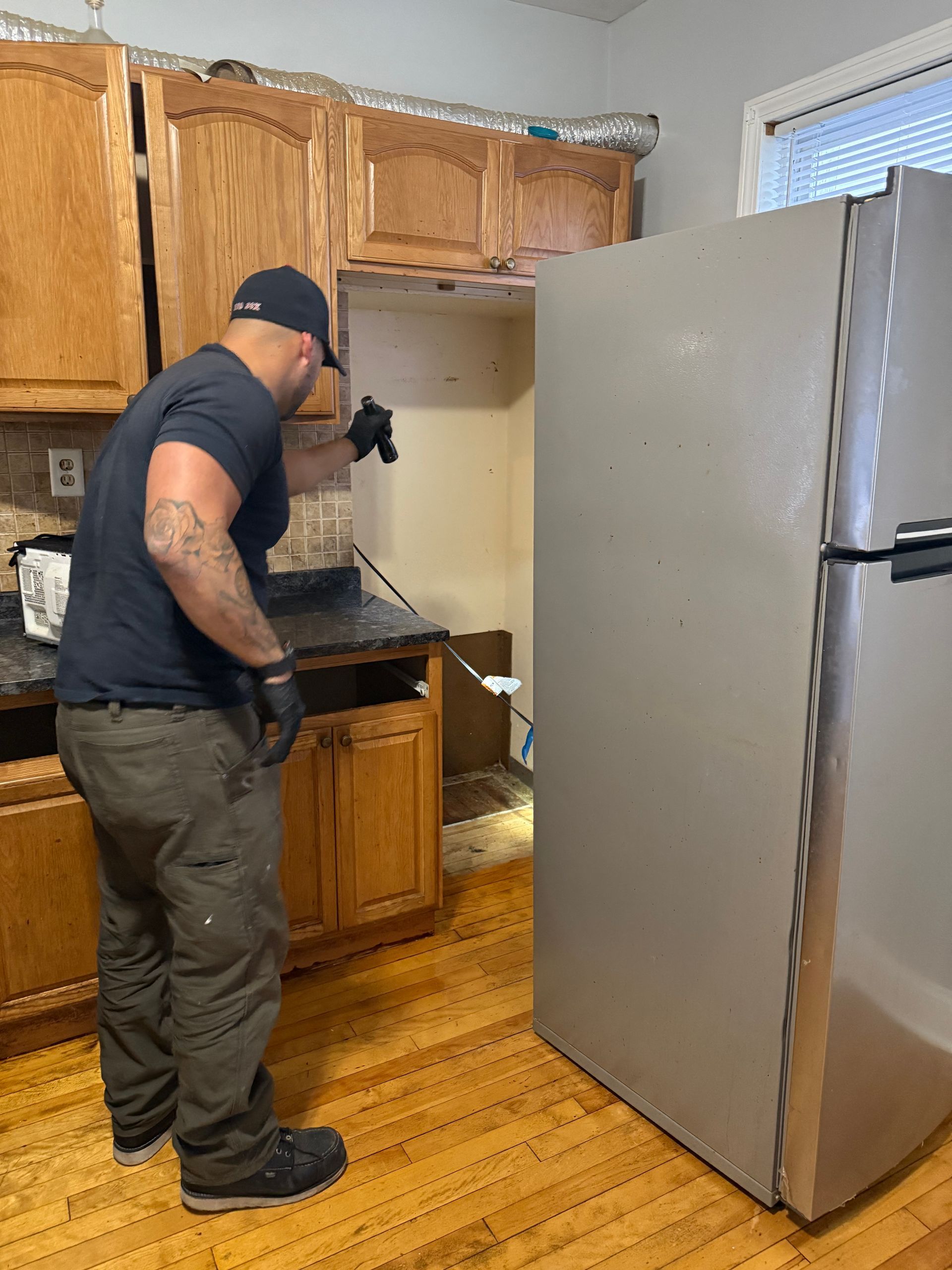 A person inspects a kitchen with removed cabinets, a refrigerator, and wood flooring.
