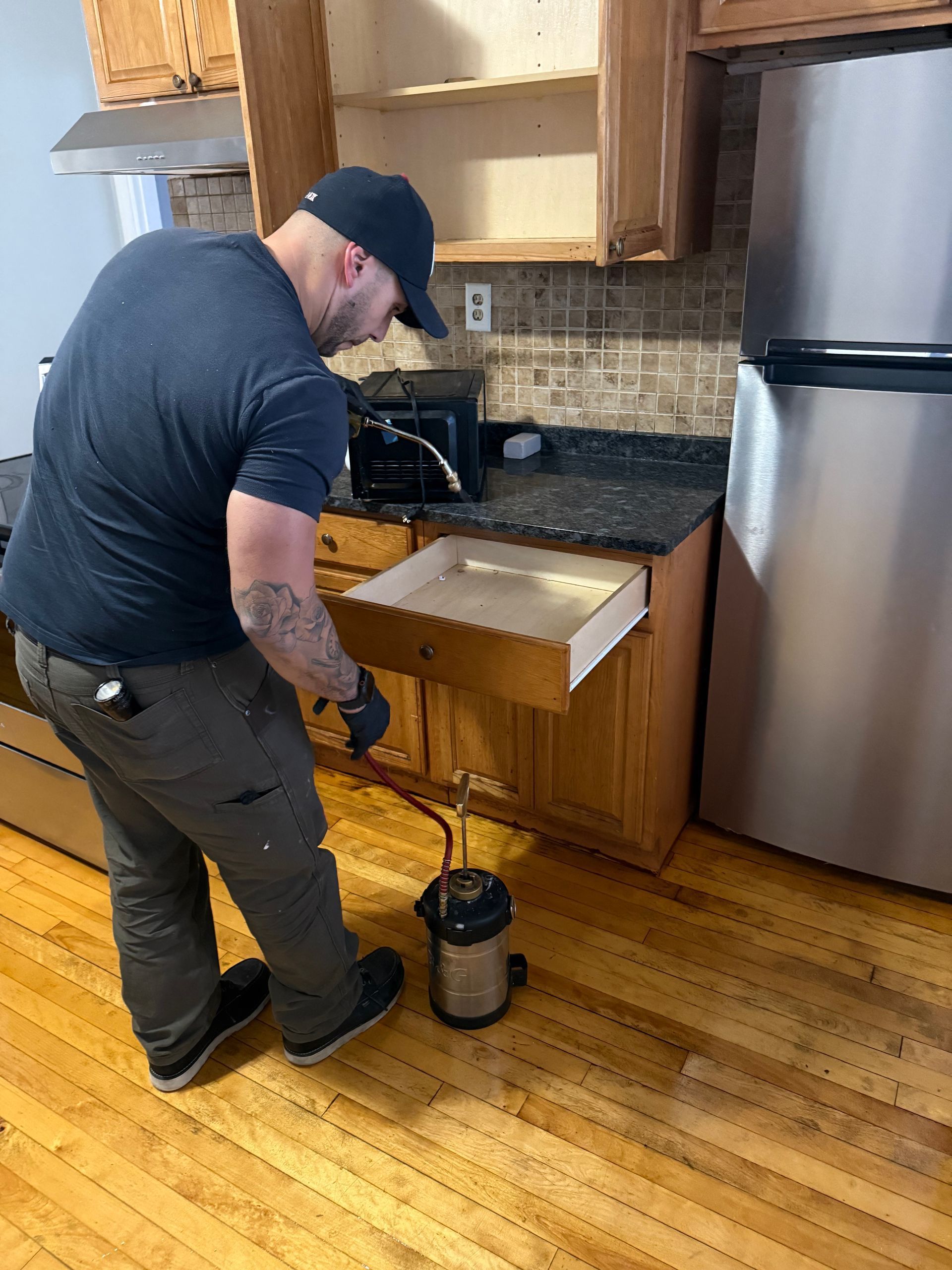 Person in kitchen, opening drawer, holding spray equipment; wood cabinets and floor.