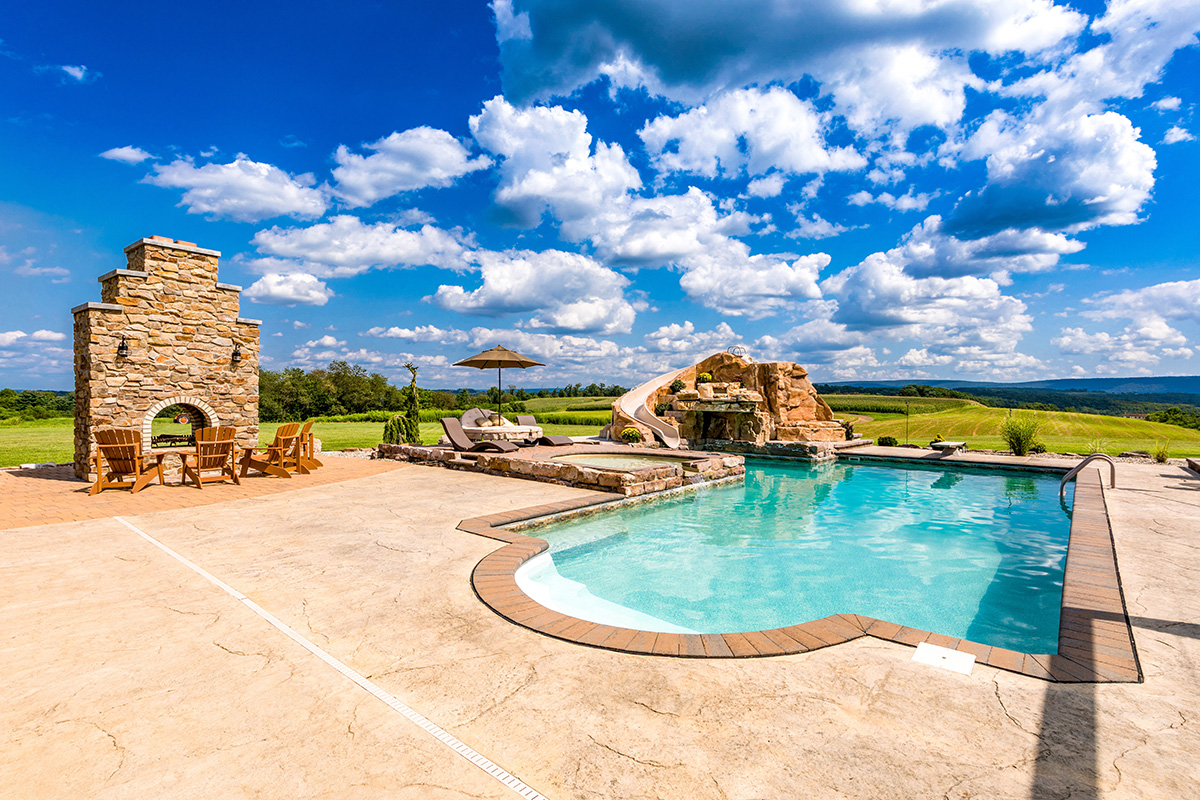 A large swimming pool surrounded by chairs and a stone fireplace on a sunny day.