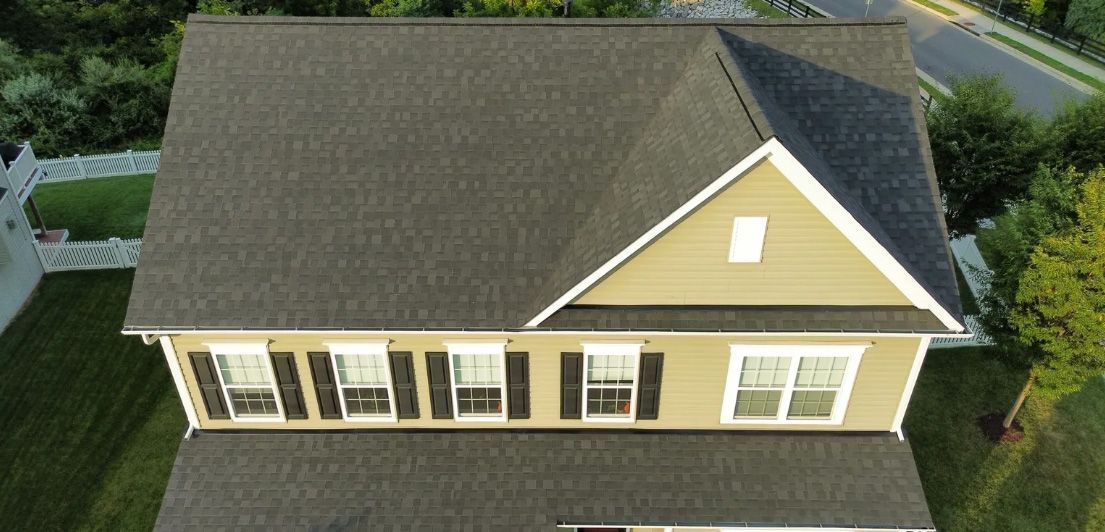 Overhead view of a house with a dark gray roof, tan siding, black shutters, and surrounding greenery.