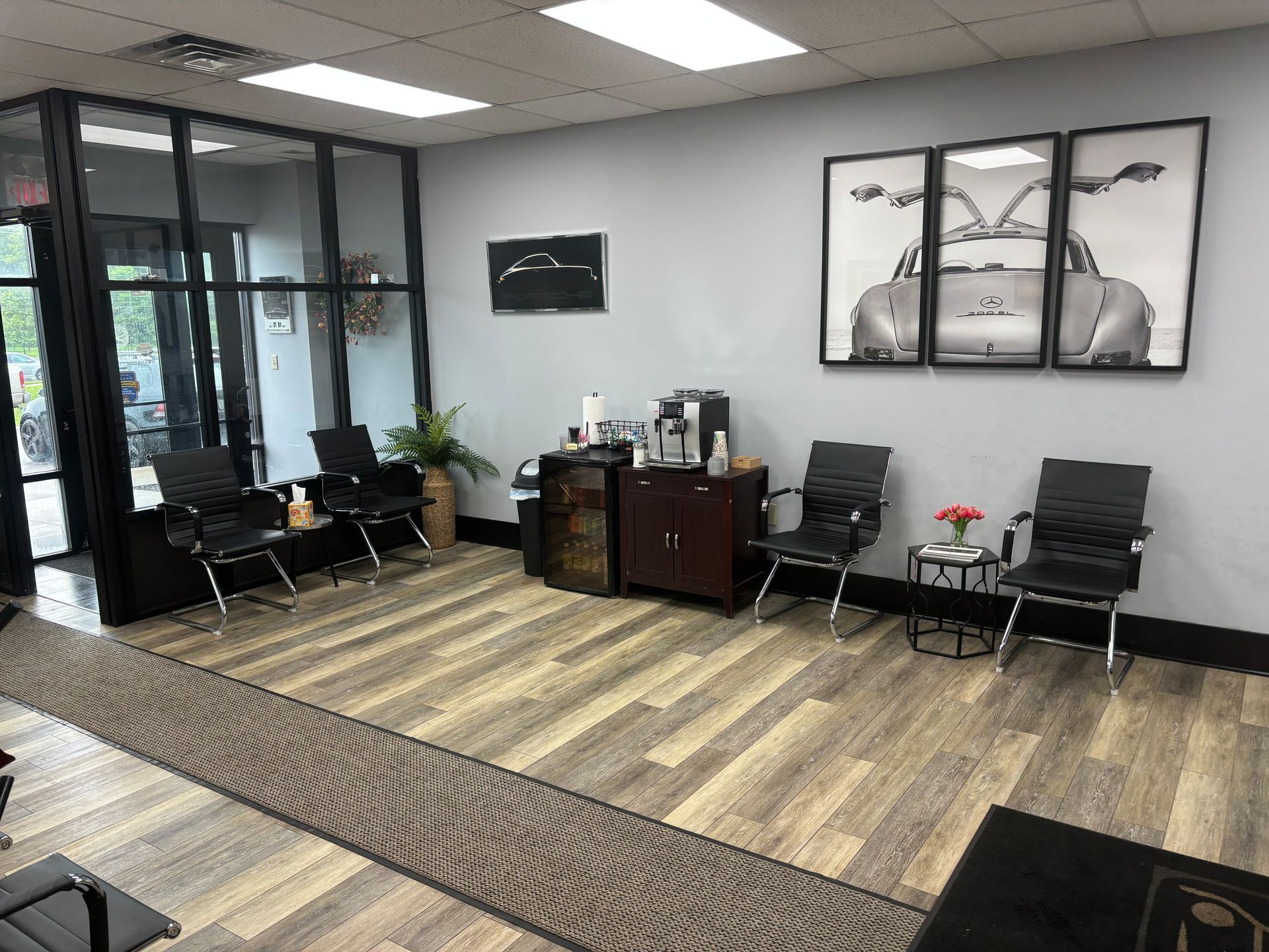 A waiting room with chairs , a coffee maker , and a picture of a car on the wall.