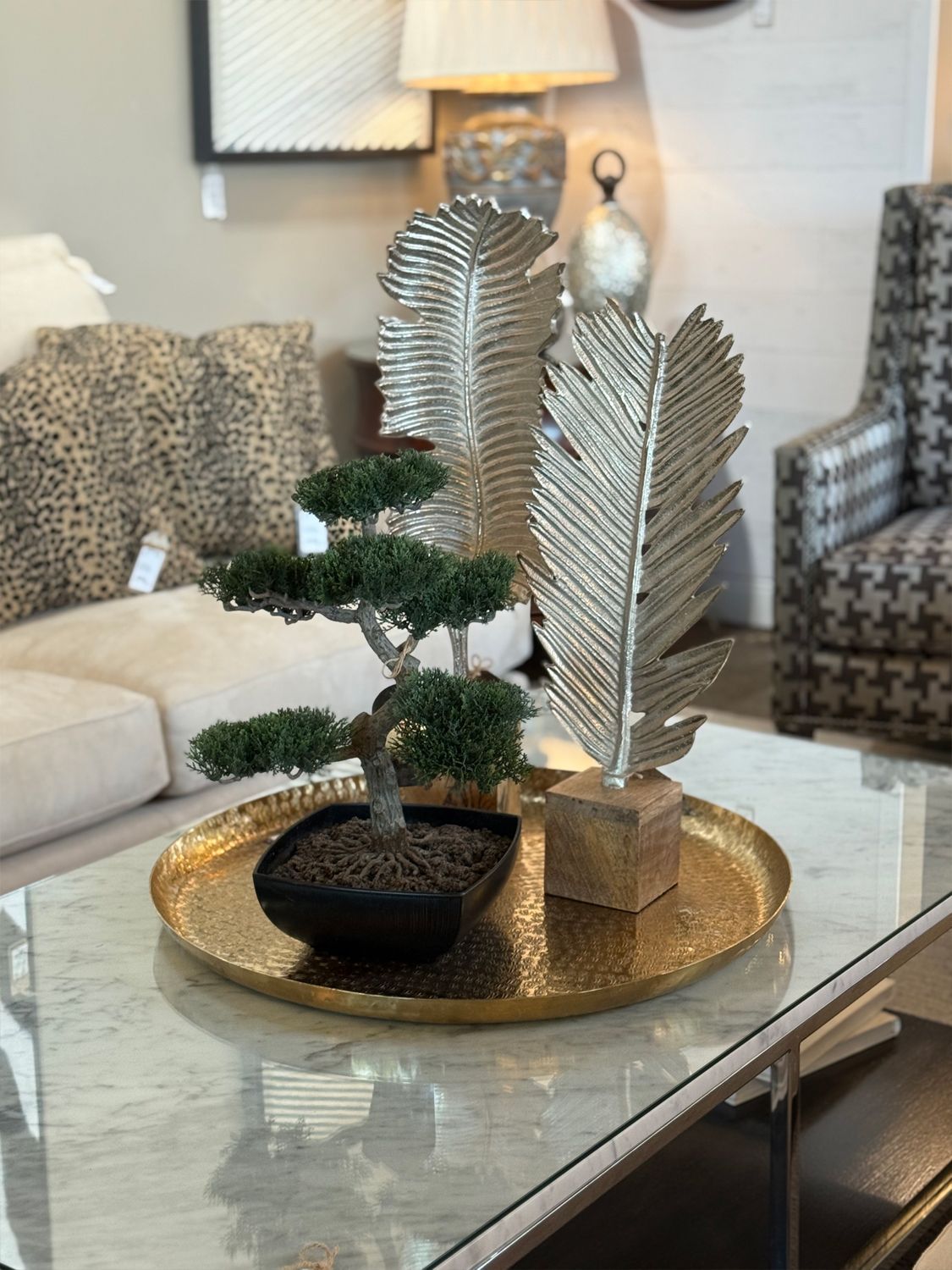 Gold tray with bonsai tree and decorative silver feathers on a glass table.