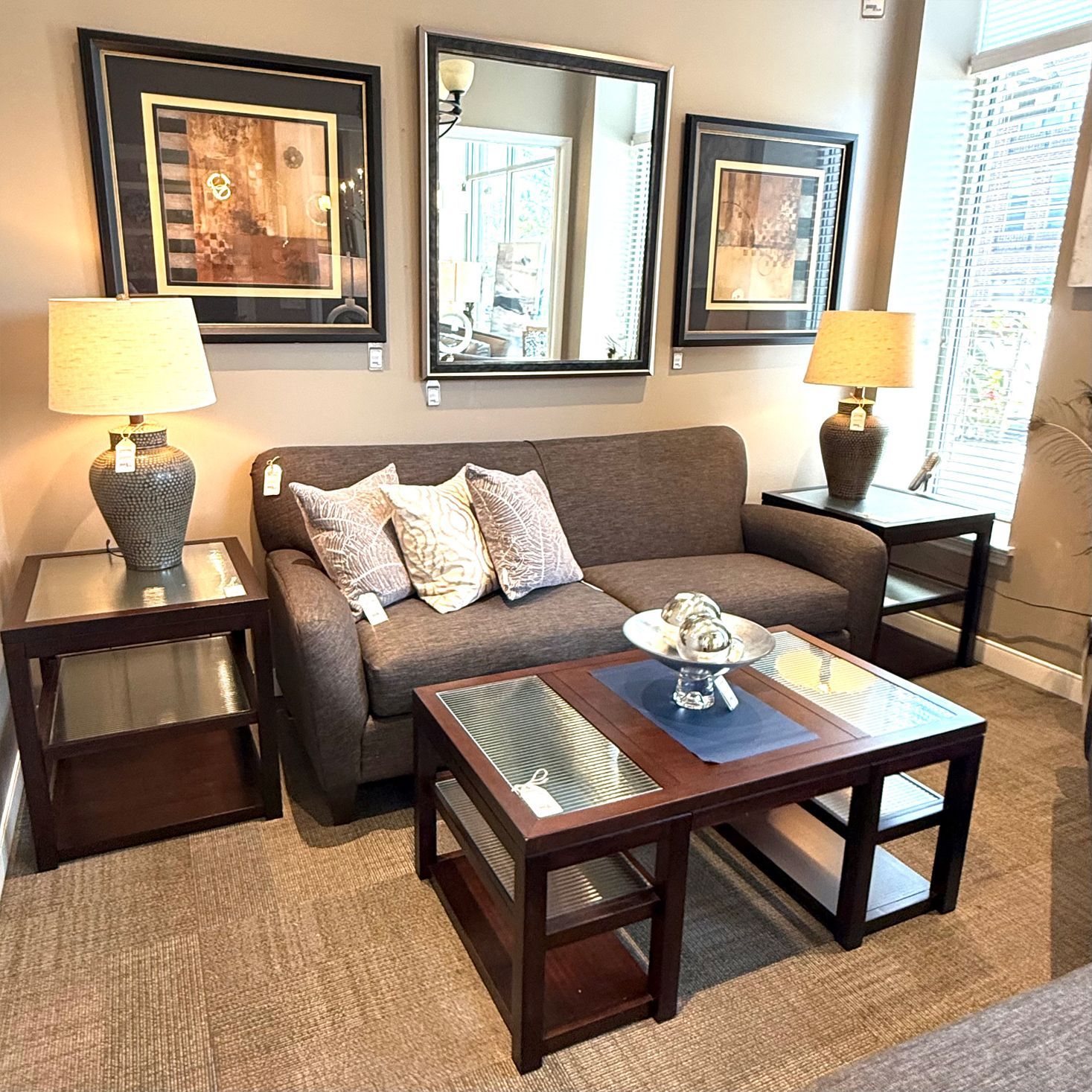 Living room furniture display: brown sofa, end tables, coffee table, artwork, mirror.