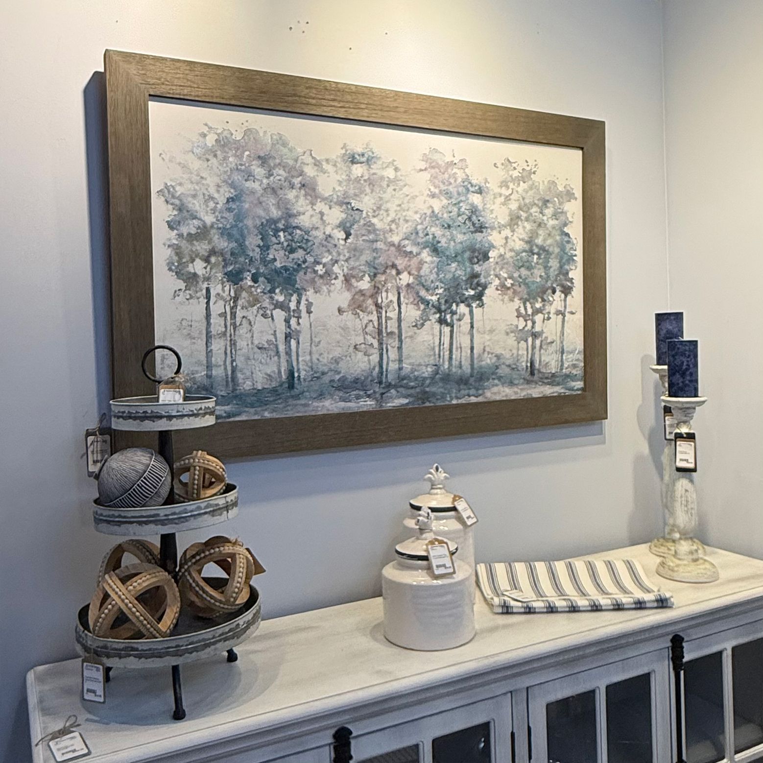 A framed painting of trees hangs above a white cabinet with decorative items like candles and orbs.
