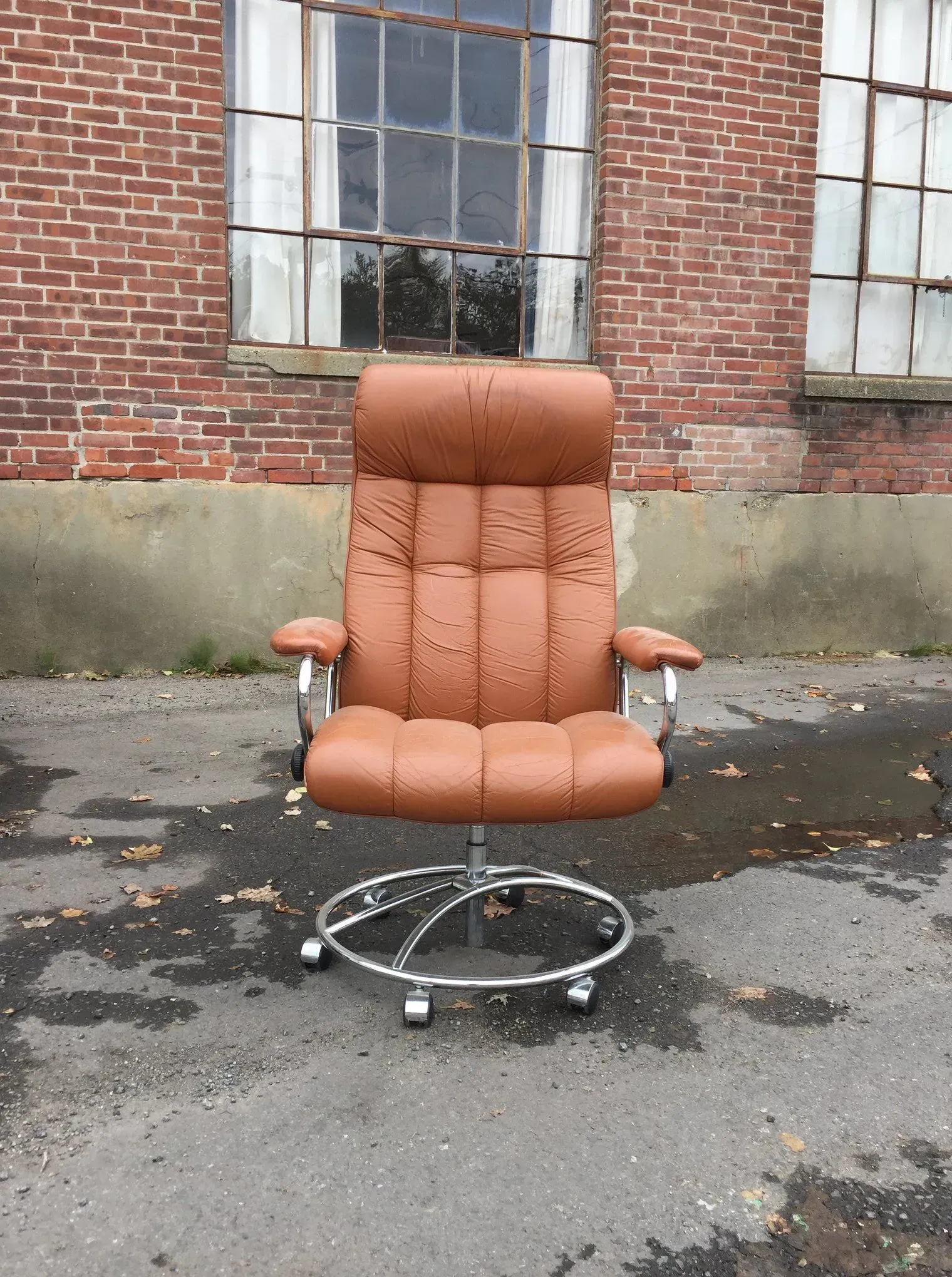 Brown office chair on a wheeled base outdoors against a brick wall.