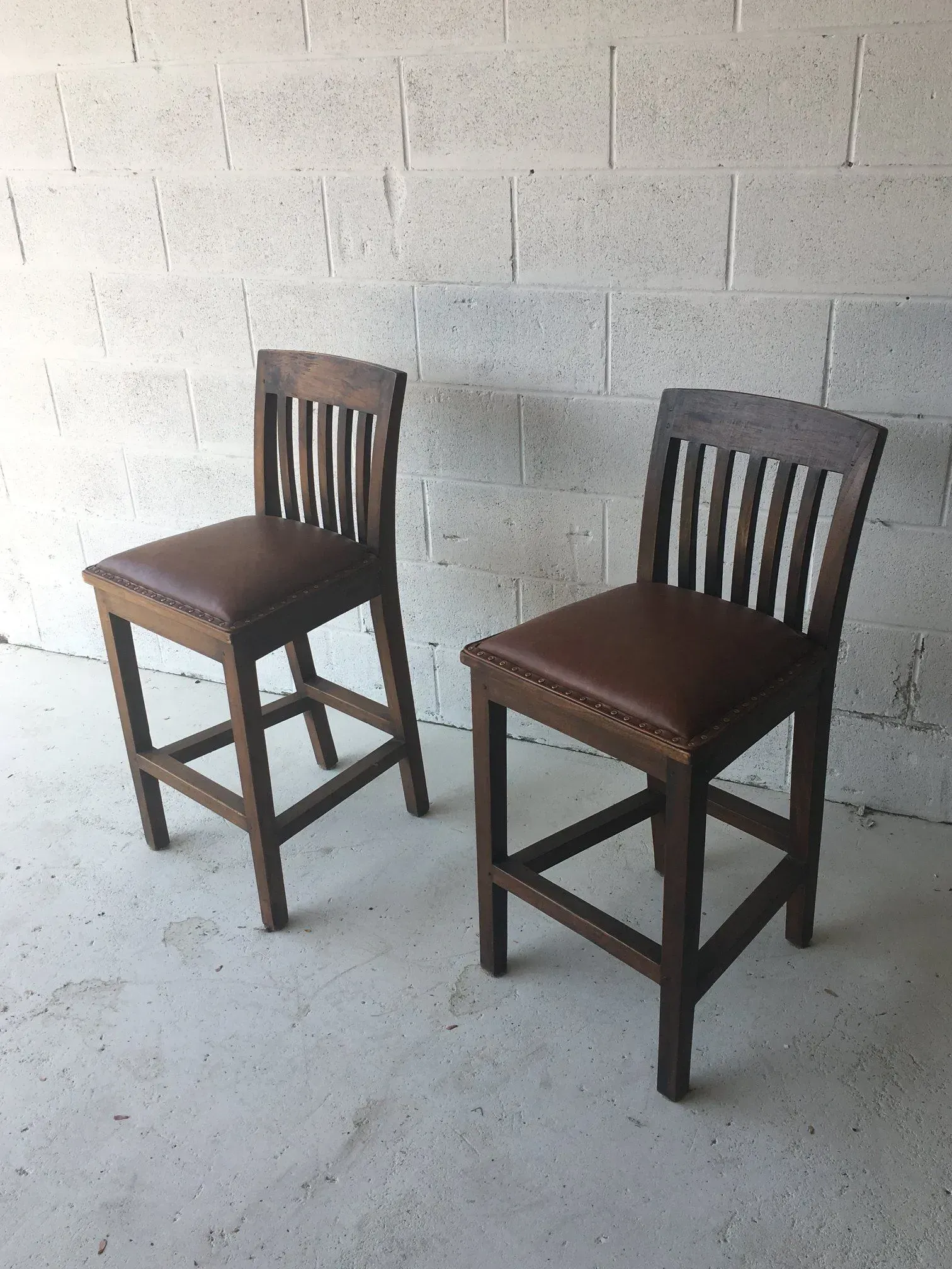 Two brown wooden chairs with slatted backs and padded seats on a concrete floor against a white wall