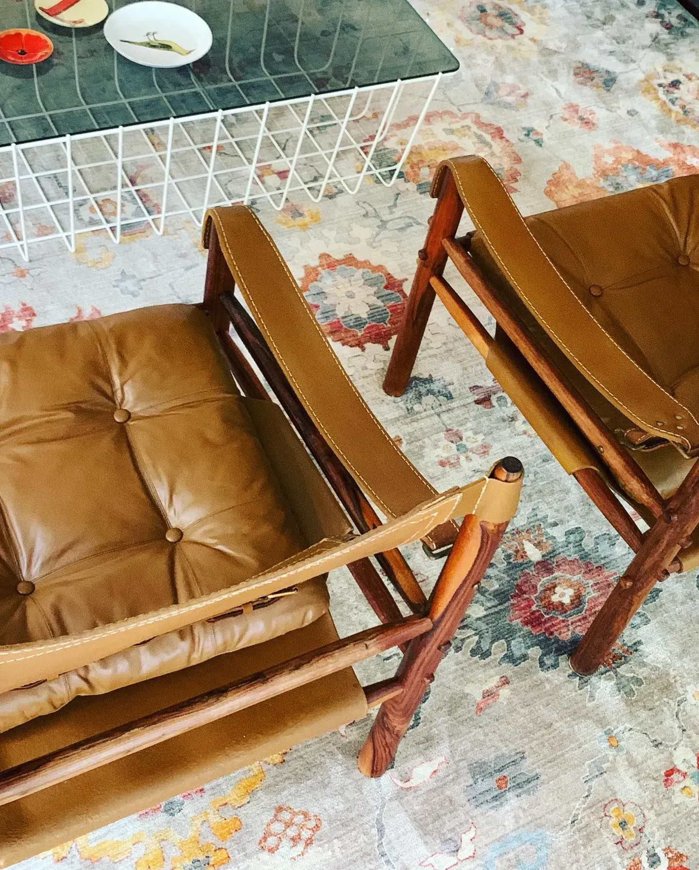 Two tan wooden lounge chairs on a floral rug beside a glass coffee table with plates and cups.