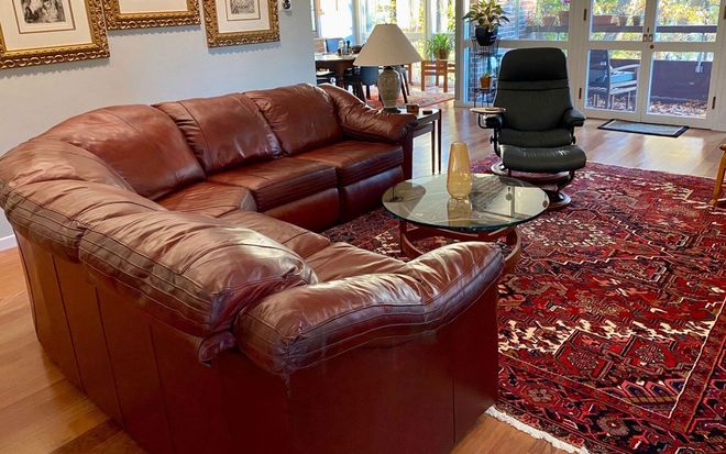Living room with a brown leather sectional, glass coffee table, black chair, and red patterned rug