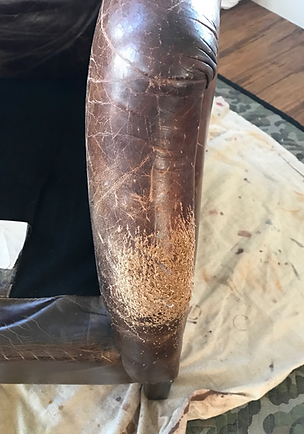 Worn brown leather chair armrest with visible cracks and scuffs beside a white cloth on a wooden floor