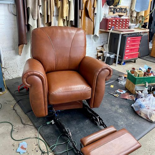 Brown leather recliner chair in a cluttered workshop, with its footrest detached in front.