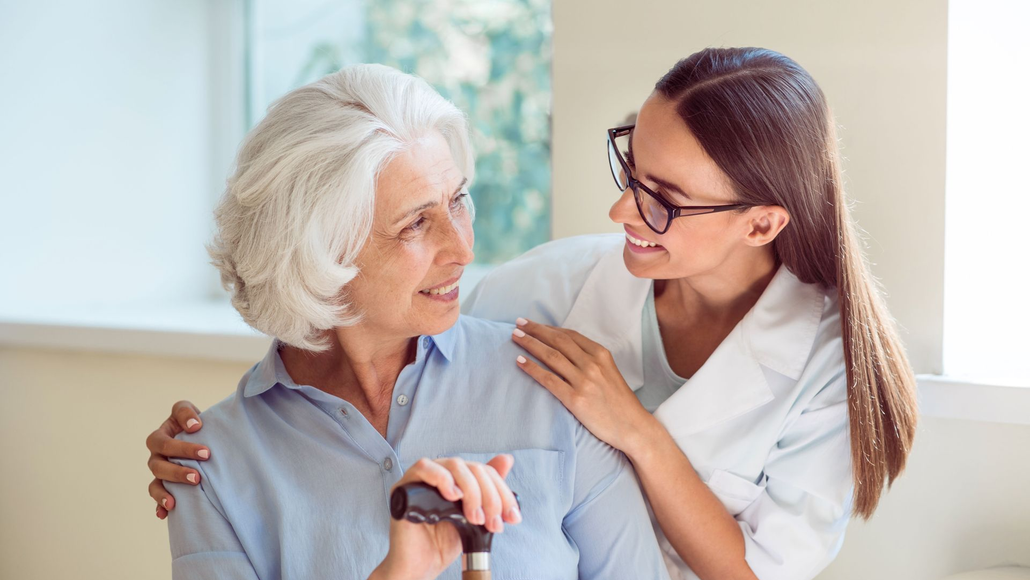 A caregiver in a white uniform places a supportive hand on the shoulder of a person holding a cane, both smiling.