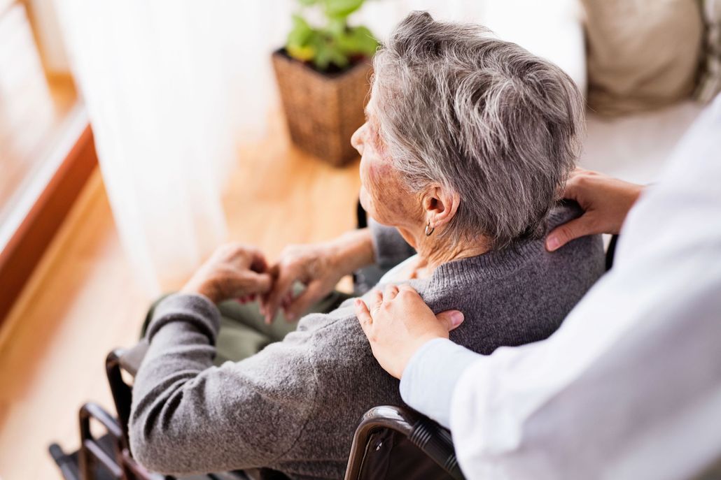 A caregiver rests a hand on the shoulder of a person seated in a wheelchair, looking out a window.