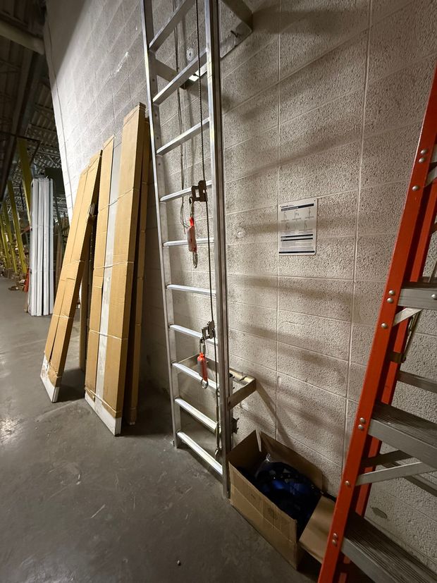 A metal ladder leans against a rough white wall, beside cardboard planks and an orange step ladder in a storage area.