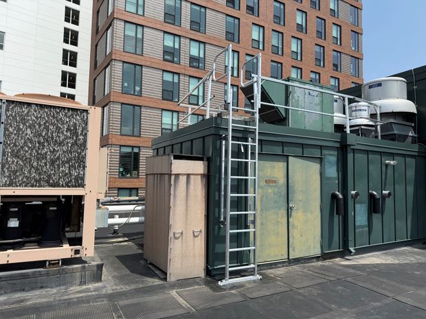 A rooftop mechanical area featuring a dark green equipment enclosure, a metal ladder, and a large air conditioning unit.