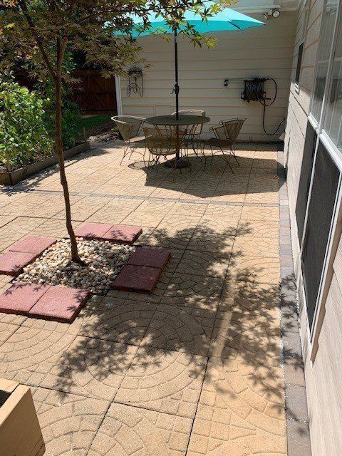 Outdoor patio with tan patterned pavers, seating area with a teal umbrella, and a small tree.