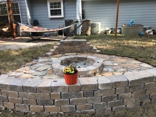 A brick fire pit with a flower pot on its wall, in a yard with a hammock and a house in the background.
