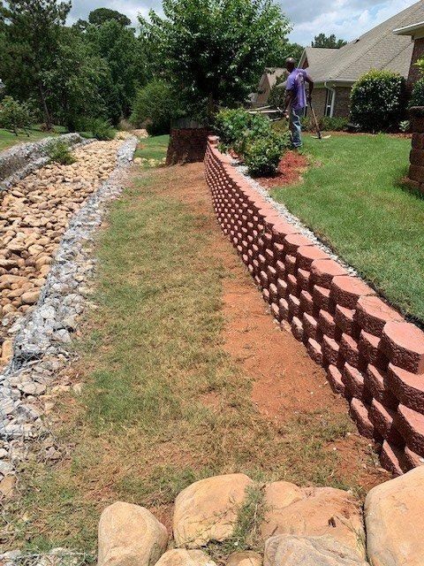 A red brick retaining wall borders a lawn. A person stands near the wall, trees, and a dry creek bed are present.
