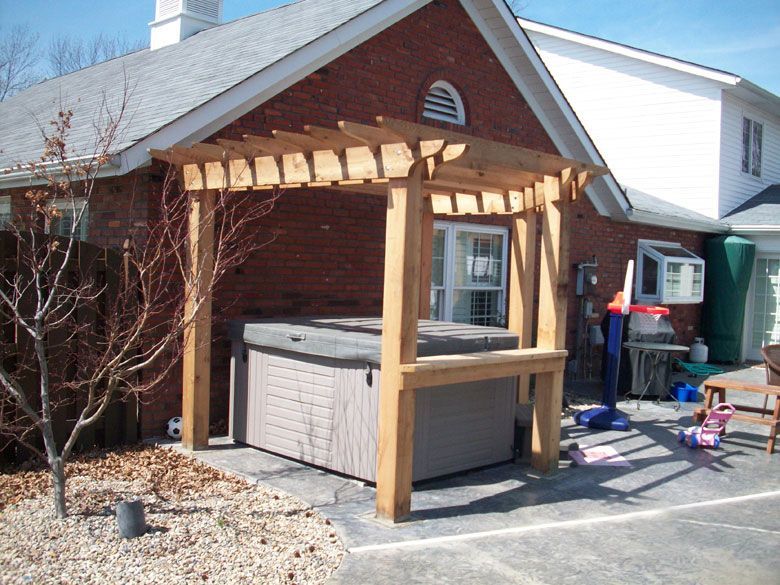 a hot tub under a wooden pergola in front of a brick house