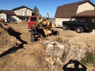 A stump grinder removing a tree stump in a yard; two trucks, houses, and dirt are visible.