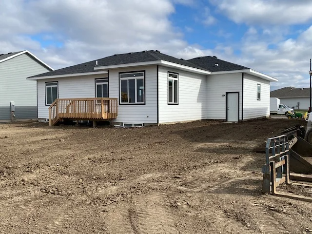 A white house with a deck is sitting on top of a dirt field.