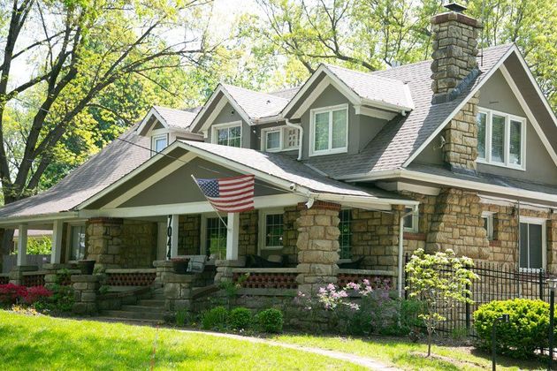 Stone cottage with green shutters and a front porch, surrounded by trees and grass, with an American flag displayed.