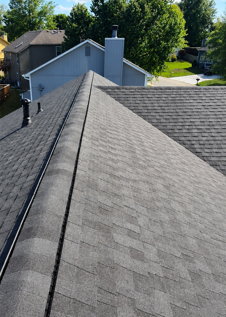 View down a gray shingled roof ridge toward neighboring houses and trees on a sunny day