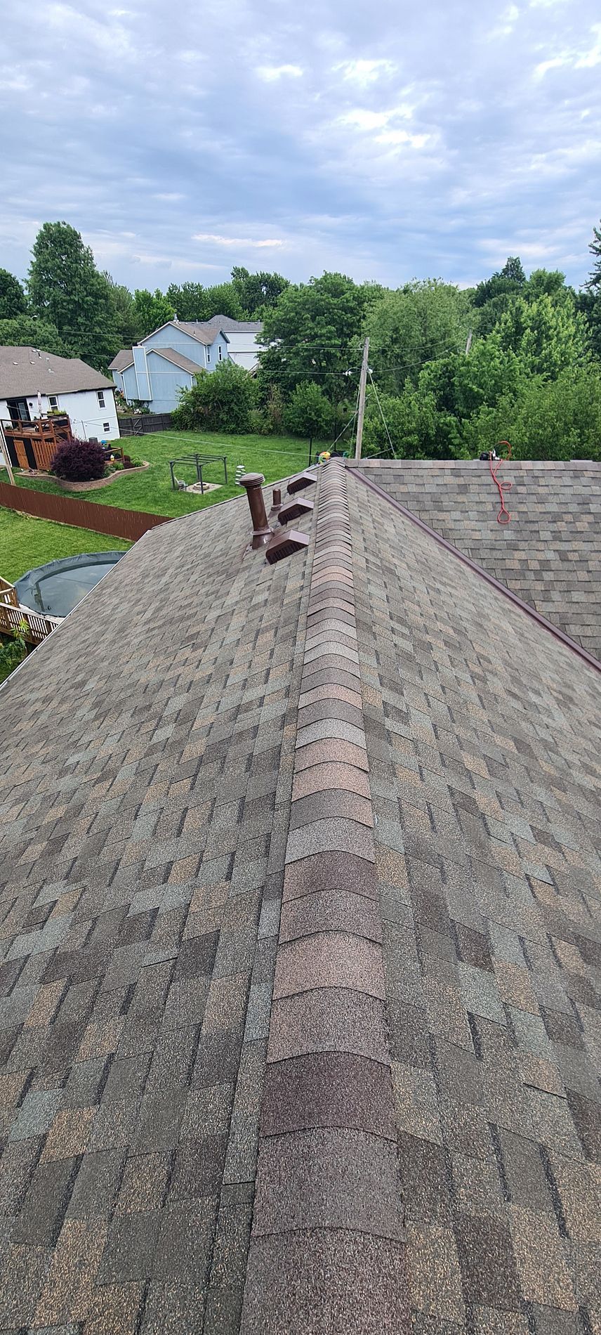 Rooftop shingles with a vent pipe, overlooking a green suburban neighborhood under cloudy sky