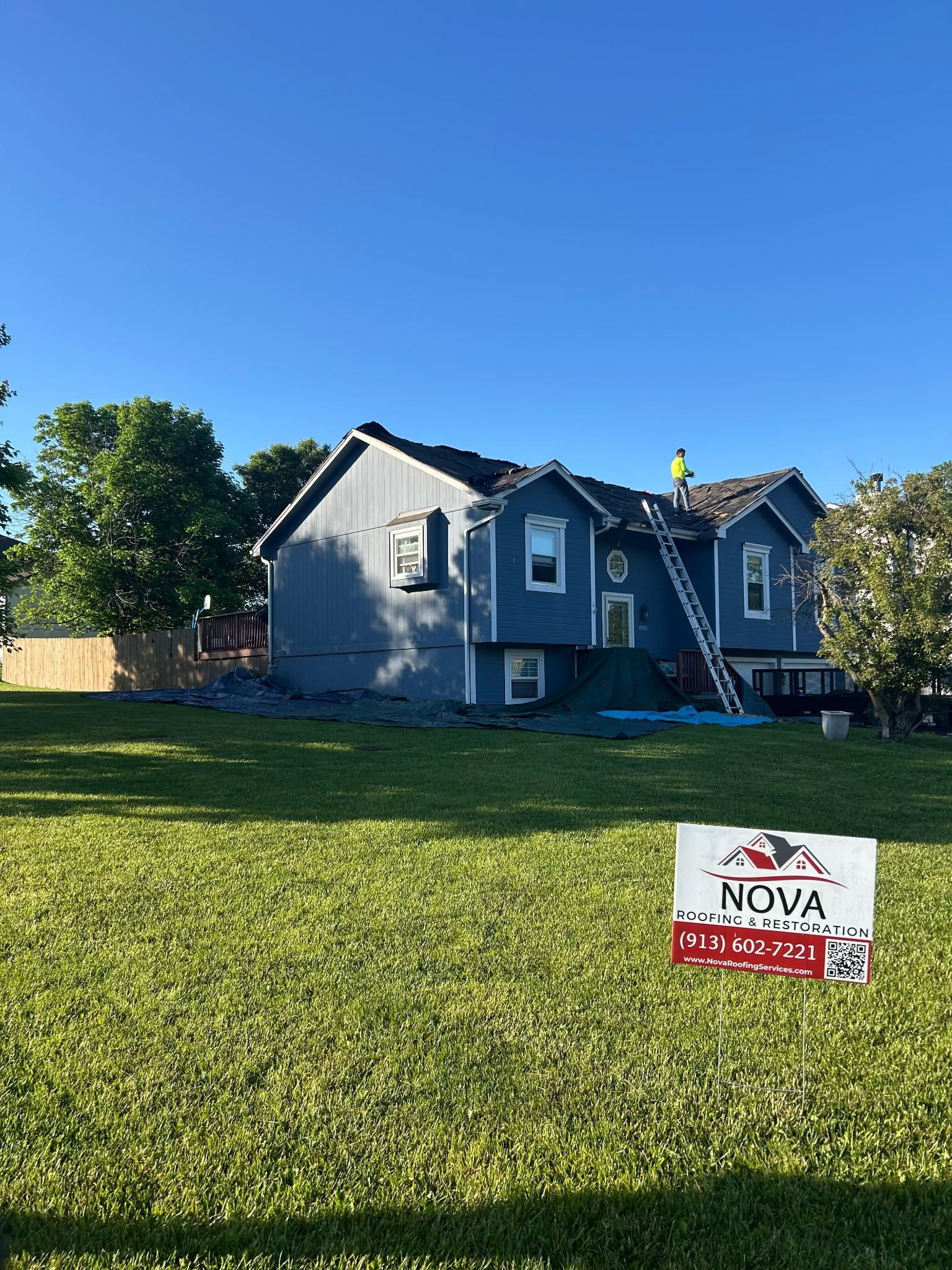 Blue house with a ladder in the yard and a NOVA sign on the lawn under a clear sky.