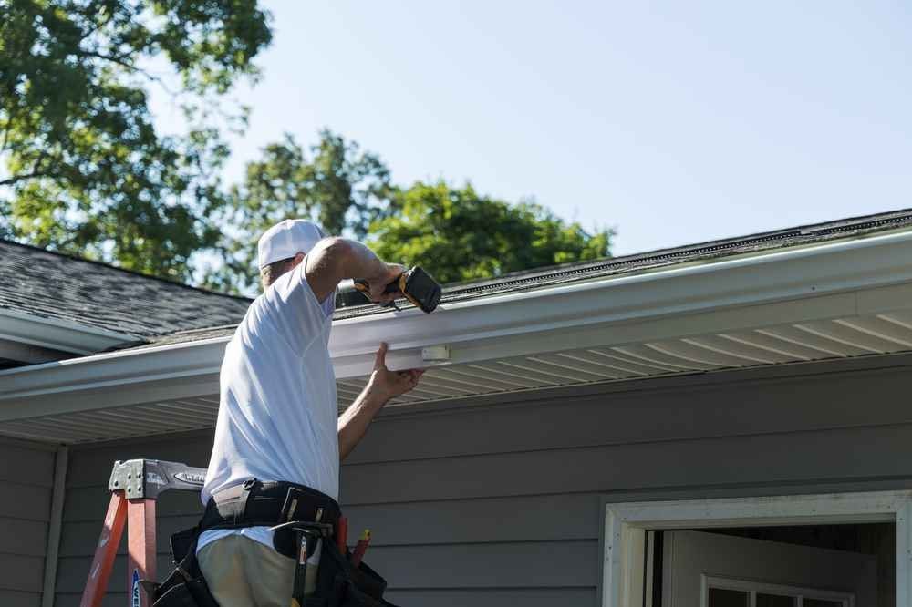 Worker cleaning gutters on a gray house roof with a ladder and power tool