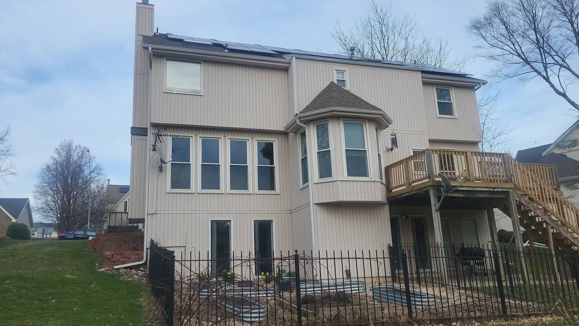 Two-story beige house with damaged siding and boarded windows, seen behind a black fence.