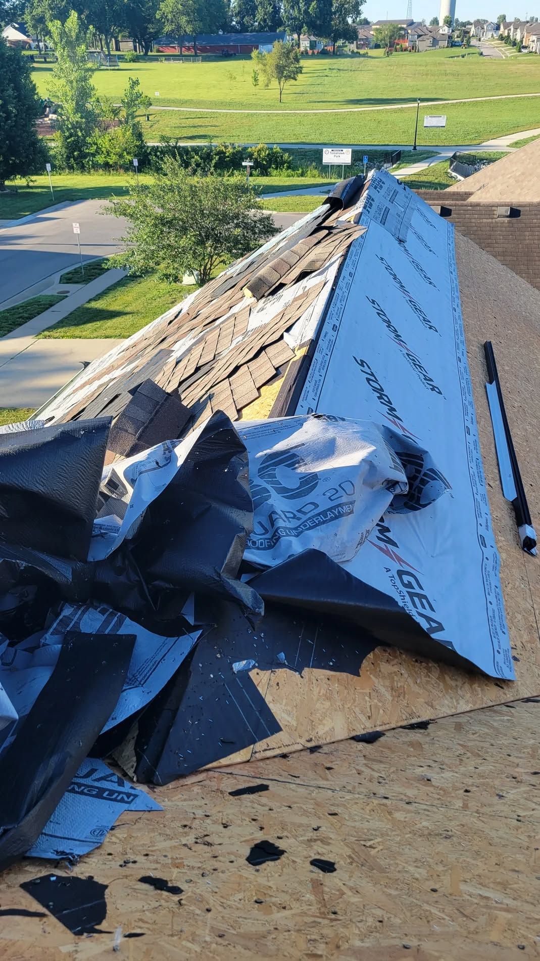 Damaged blue roof shingles and debris on a rooftop, with green fields in the background