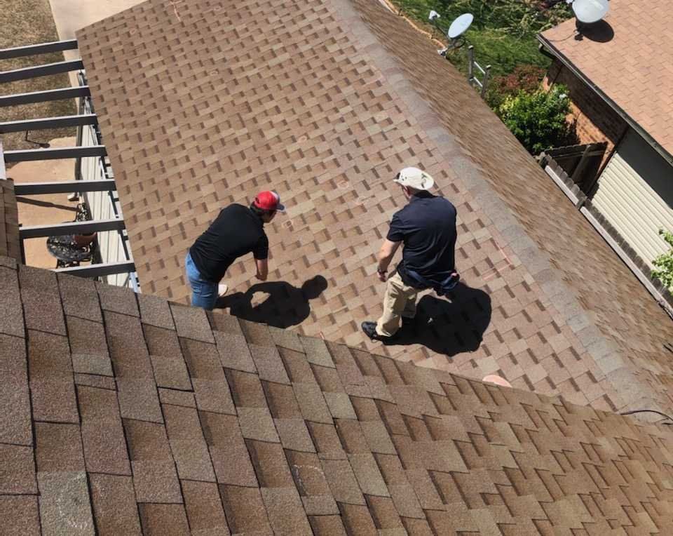 Two workers inspecting a shingled roof, one crouched and one standing with tools.