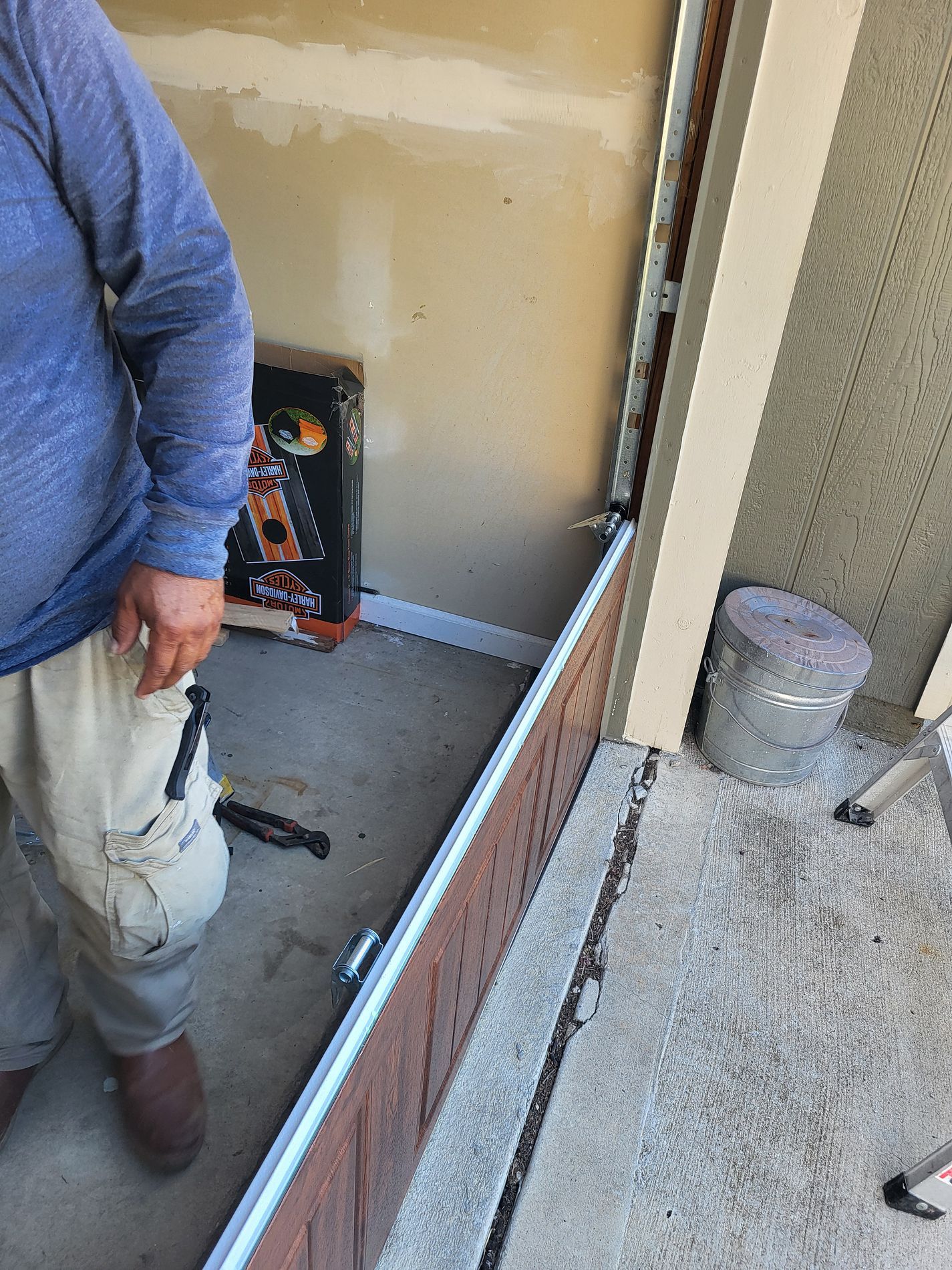 Worker in blue shirt standing in a narrow unfinished room with tools and a trash bin nearby.