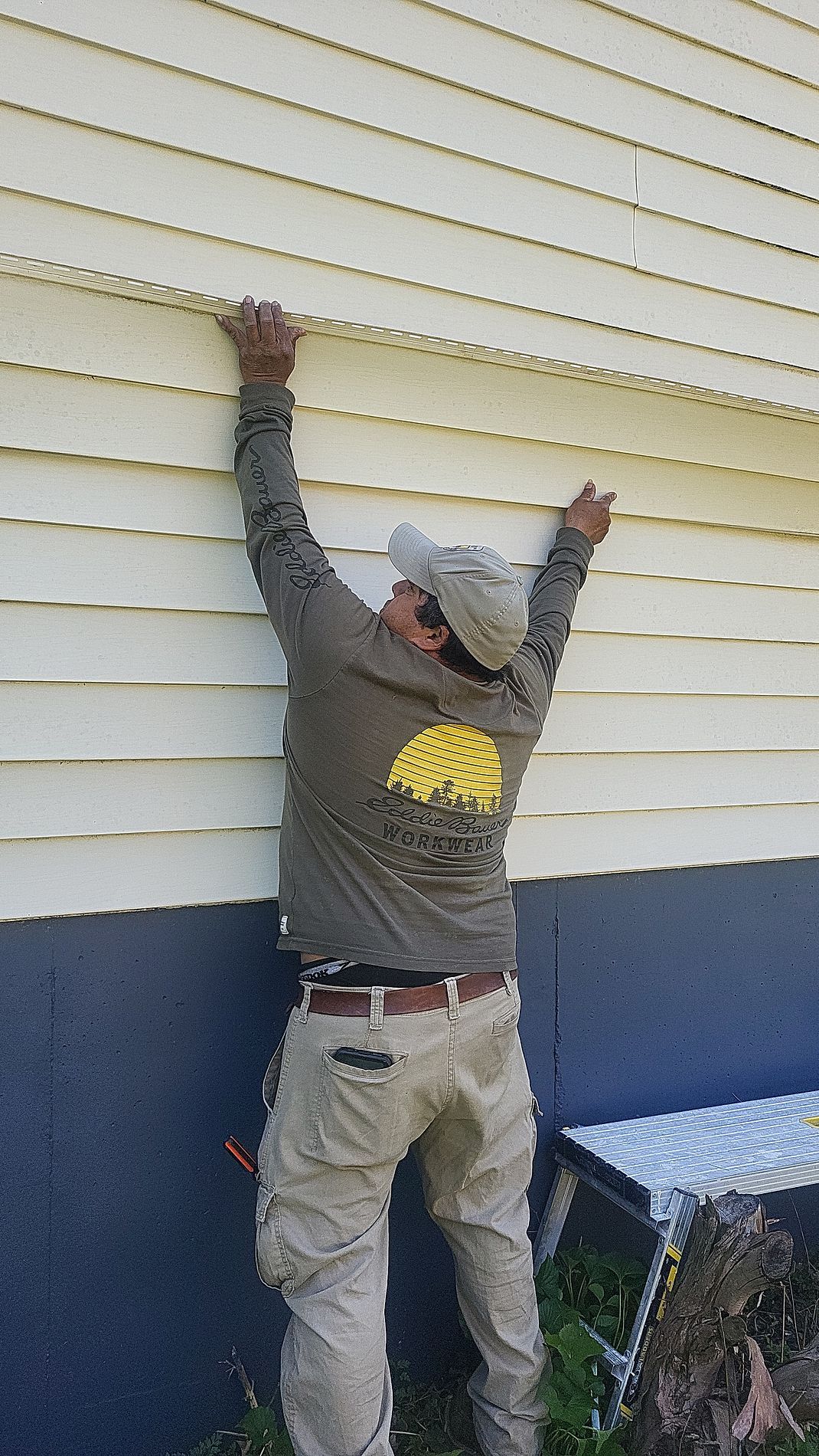 Person in a gray sweatshirt and cap reaches up to work on cream-colored siding outside a building.