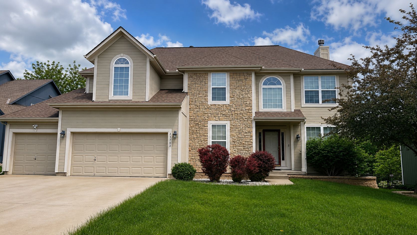 Two-story suburban house with beige siding, three-car garage, and a green lawn under a partly cloudy sky
