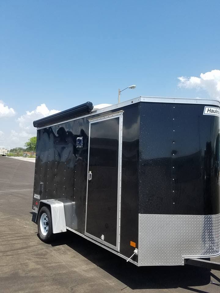 Black enclosed cargo trailer with silver trim parked on a paved lot under a blue sky