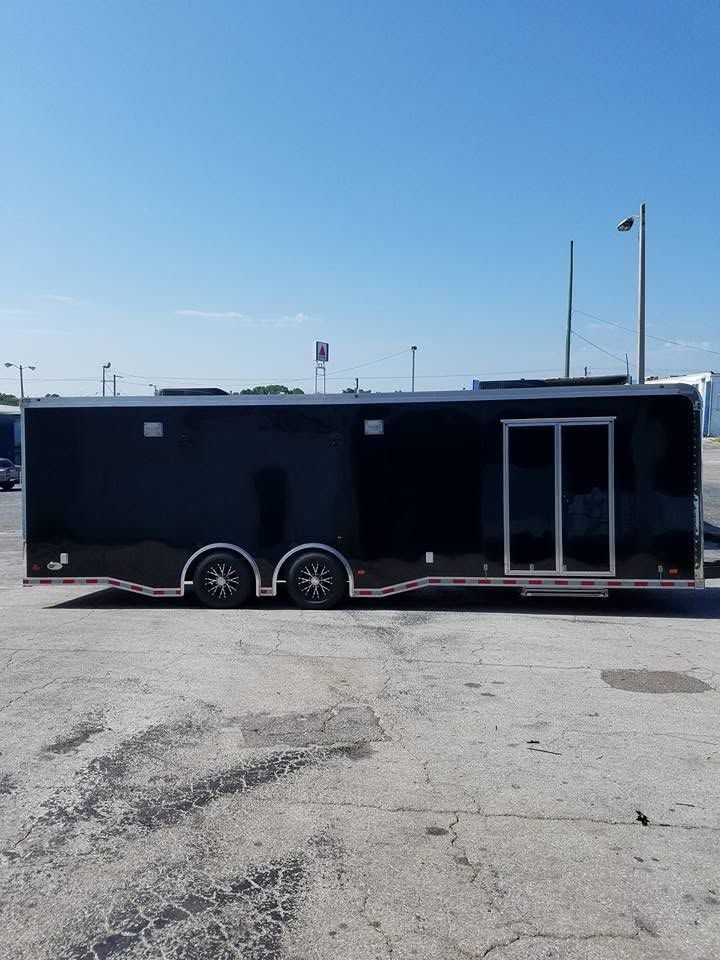 Black enclosed trailer with dual wheels parked on a sunny paved lot under a blue sky