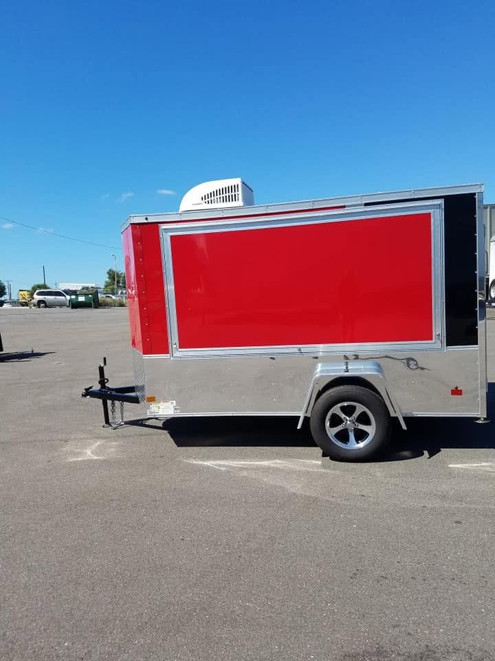 Small silver cargo trailer with a bright red side panel parked on an open asphalt lot.