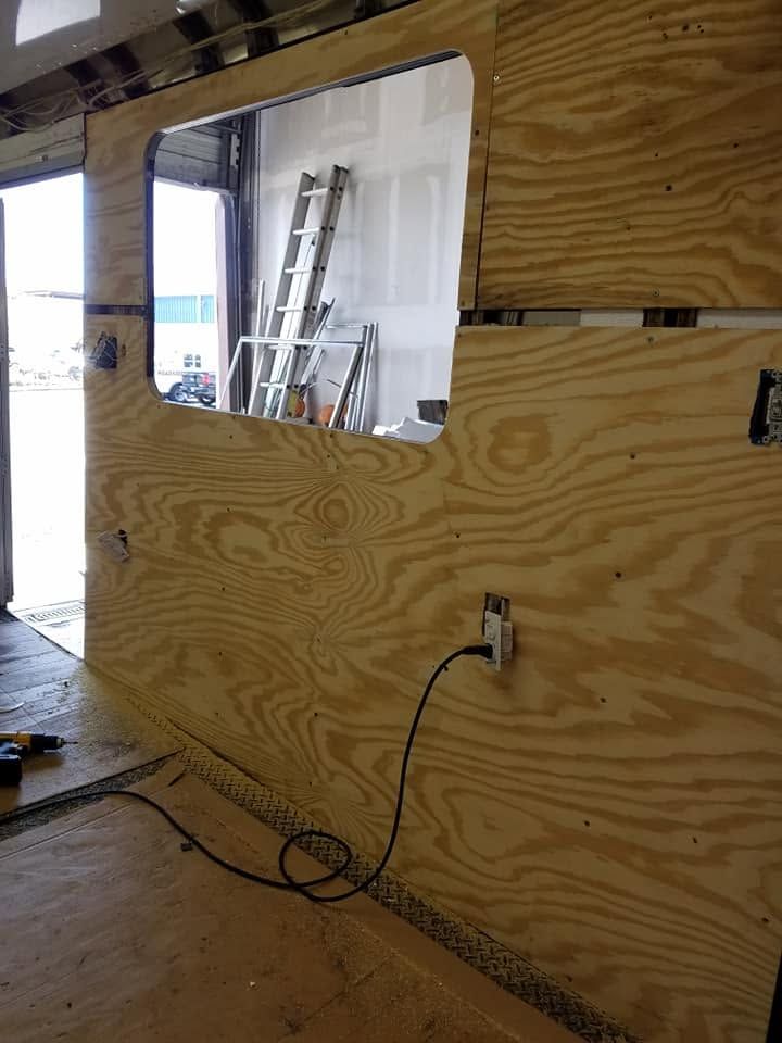 Plywood barrier with a window opening inside a construction area, with a power outlet and cord on the wall.